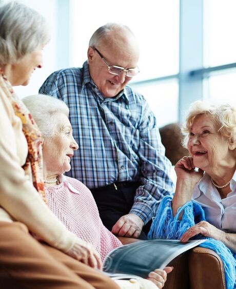 Vier ältere Menschen sitzen in hellem Raum; drei unterhalten sich lachend mit Frau in blauer Stola mit Fotoalbum.