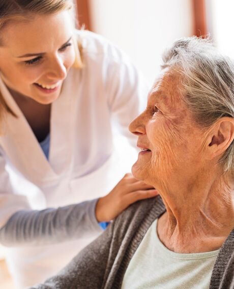 Jüngere Pflegekraft im weißen Kittel lächelt ältere Frau an und legt fürsorglich die Hand auf ihre Schulter.